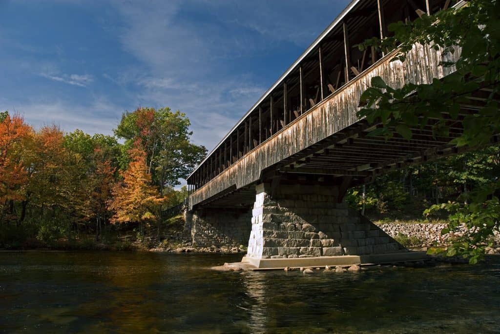 A wooden covered bridge over water has trees with autumn colors surrounding it under blue skies