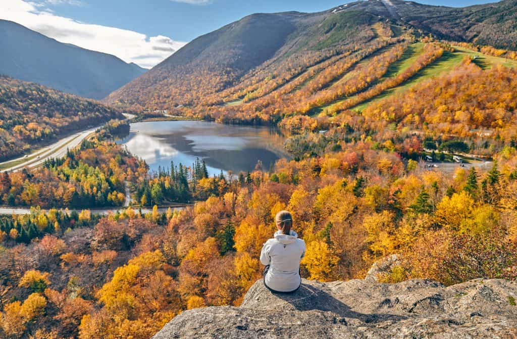 Someone sitting on a rocky ledge looking at a view of water and mountains with fall leaves