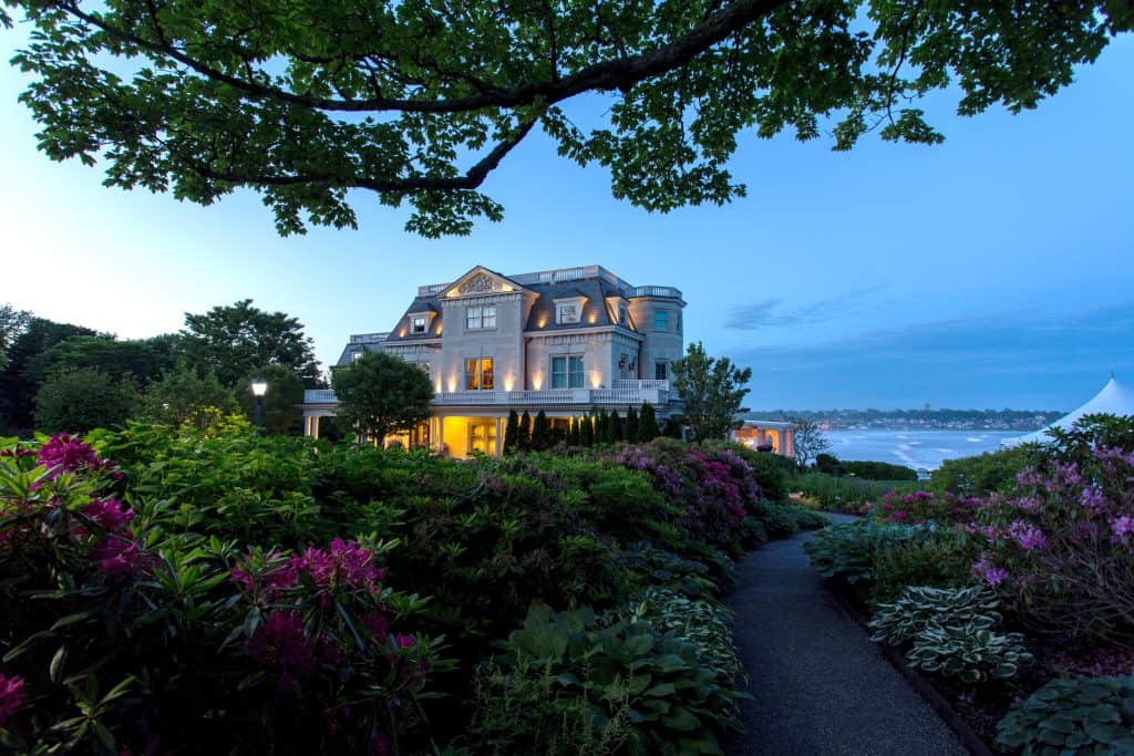 Romantic New England building is lit up in the evening by lights. In the foreground is a garden with a path leading to the home. The background has a view of the ocean.
