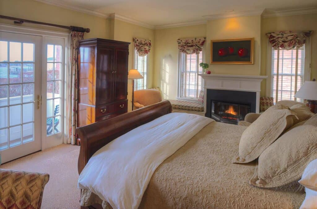 Interior of a bedroom in a hotel. The bed is plush and is made with a tan comforter. Behind the bed is a fireplace on with artwork above the mantle.