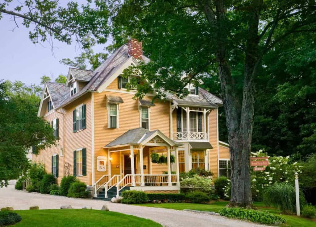 Yellow New England bed and breakfast sits with trees and green grass surrounding it.