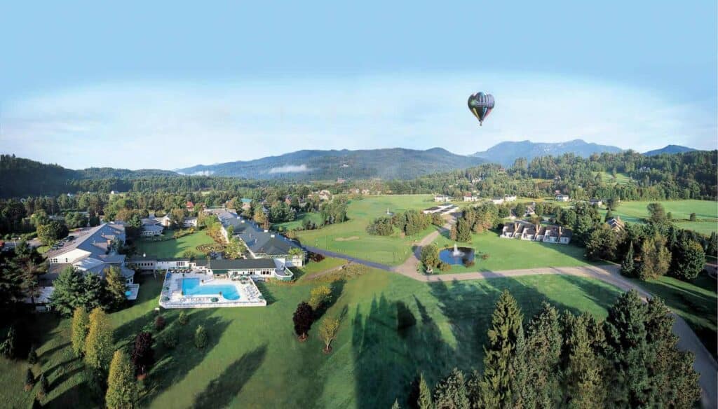 Panoramic view of a small town with mountains in the background under a blue sky. A hot air balloon is floating around the town.