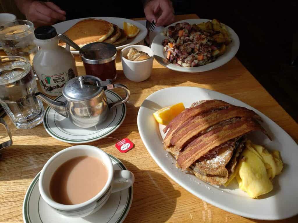 Spread of breakfast on a wooden table at a restaurant; plates contain a mixture of eggs, pancakes, toast, bacon, and other breakfast classics.