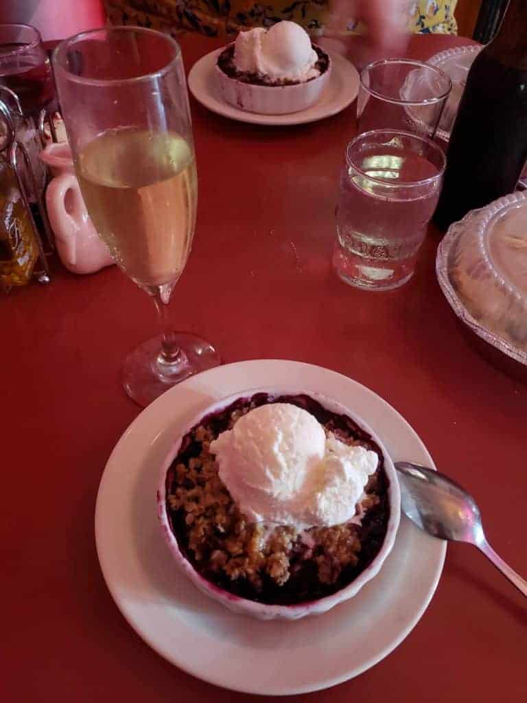 Dessert covered in white ice cream in a bowl with a spoon next to it. A glass of white wine is next to the dessert, which sits on a red tablecloth.