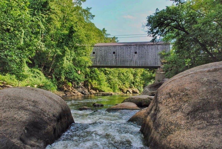 River rushing between two sides of a forest. A wooden covered bridge crosses over the river.
