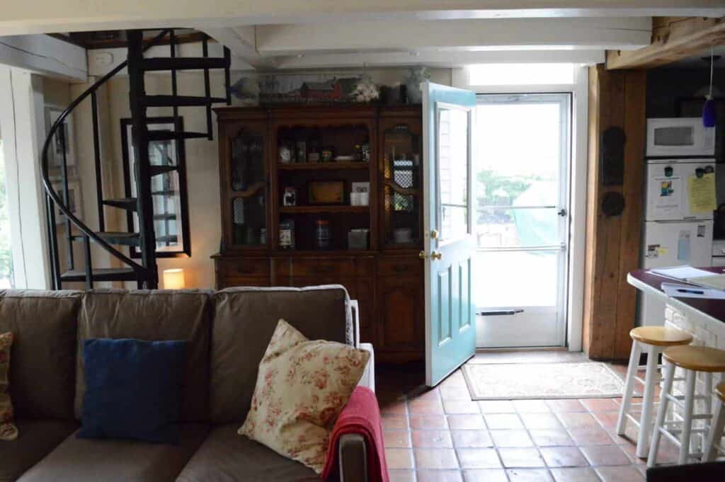 Interior of a building with the door open. A grey couch sits with a red blanket and floral pillow.