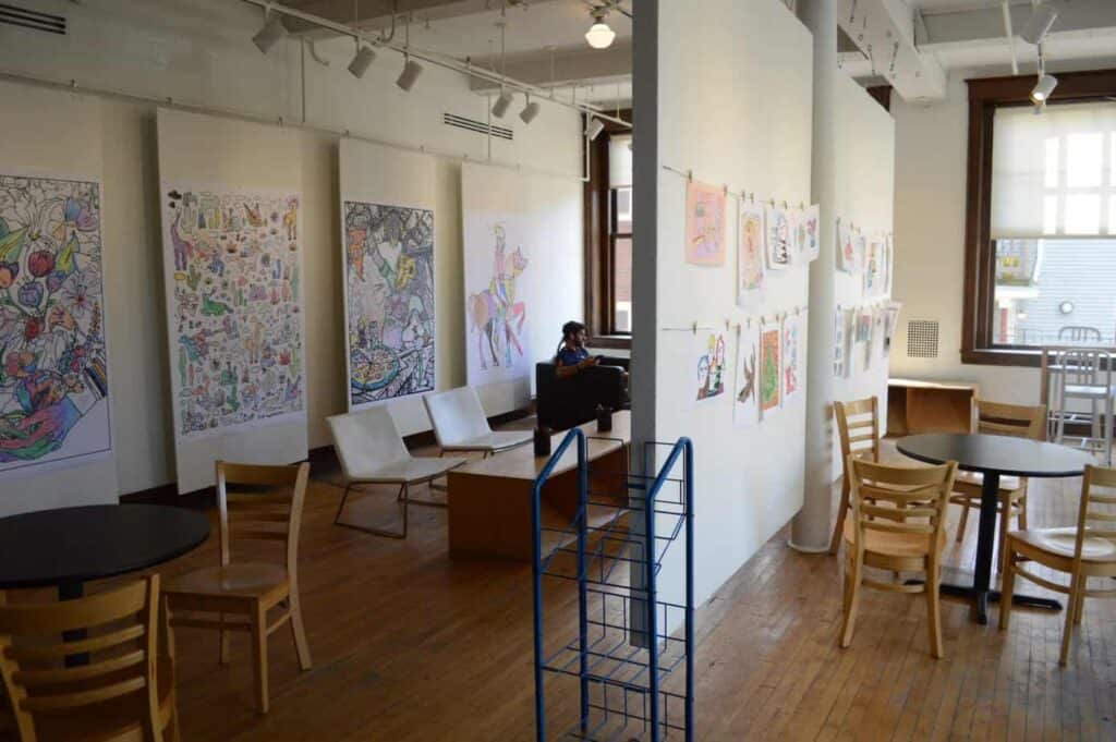 Inside a clean, white cafe with art hanging on the walls with clothespins. Cream chairs sit around tables.