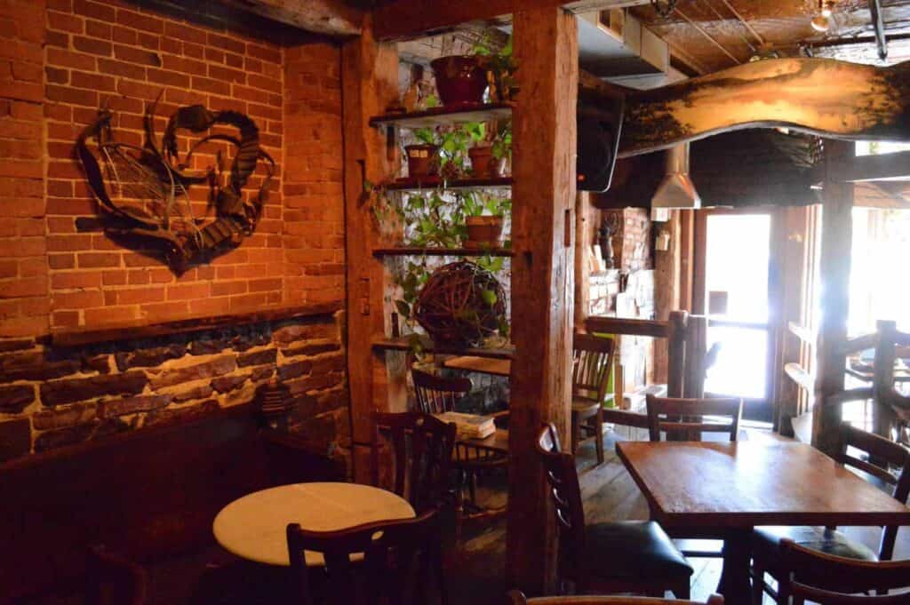 Interior of a warm coffee shop with brick walls and lots of plants on bookshelves. In the forefront, two tables are surrounded by chairs.