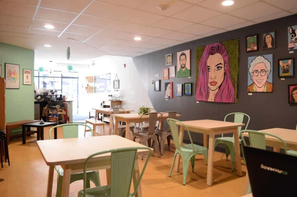 Interior of a cafe with pop art on the wall. Light wood tables are found inside surrounded by green metal chairs.