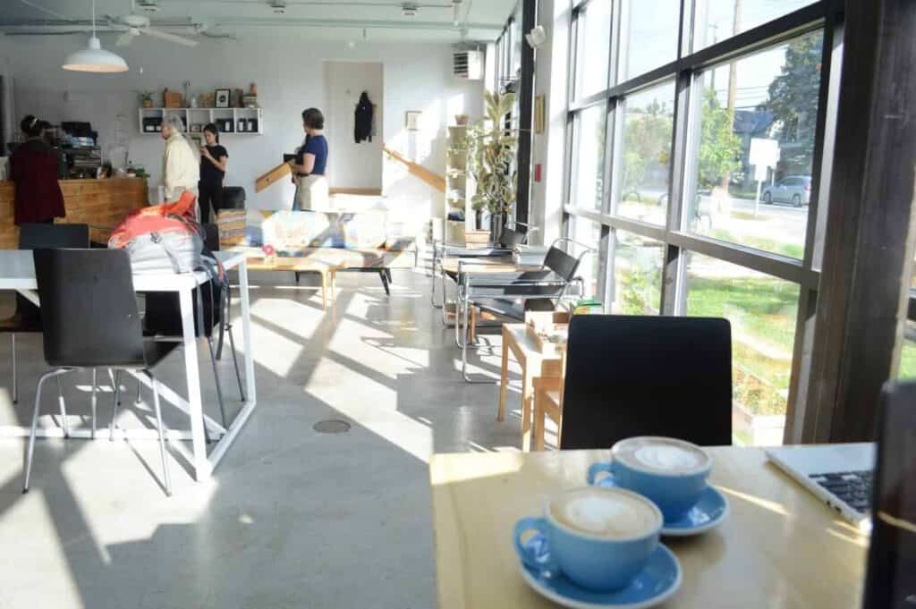 Inside of a Burlington, Vermont cafe with white walls and large windows. In the forefront, two black mugs sit on a wooden table filled with coffee. In the distance, two people stand.
