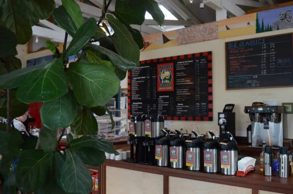 Coffee counter in a cafe with coffee lining the counter. Behind the counter is a black menu with white writing and a black and red checkerboard design lining the menu.