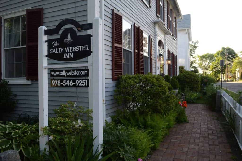 Exterior of a building that\'s blue with dark brown window detailing. A brown sign is in front of the building that says \"The Sally Webster Inn\" in white.