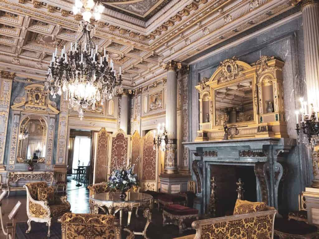 An ornate old fashioned room with a large chandelier, a fireplace, furniture and very ornate ceiling