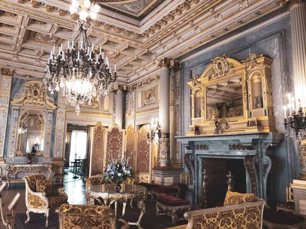 An ornate old fashioned room with a large chandelier, a fireplace, furniture and very ornate ceiling - interior of newport mansion