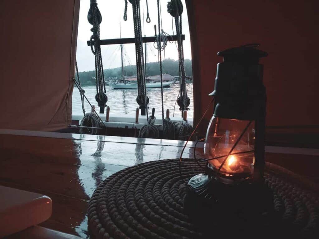 A lit lantern sits on top of a wooden table. A window is seen behind the lantern with a view of a boat floating on water.