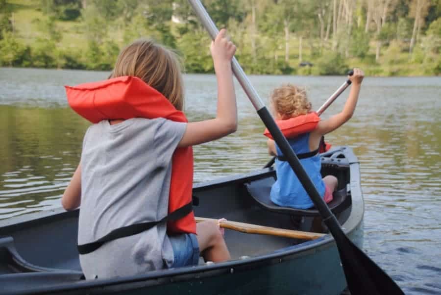 Two children with orange lifevests paddle a canoe on a lake. A forest can be seen in the background.