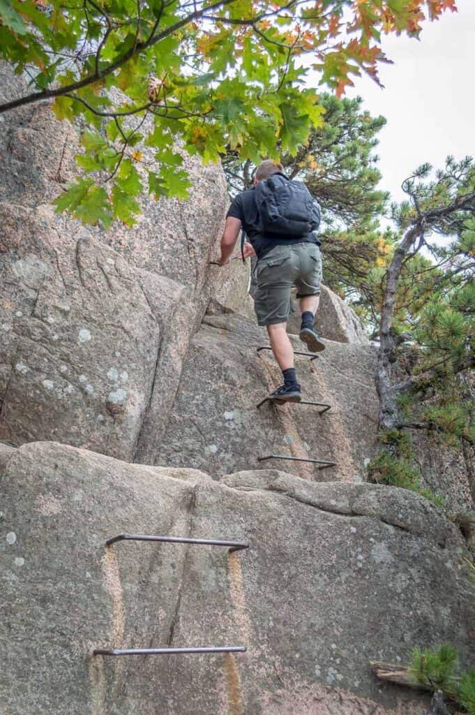 Man climbing up a metal ladder attached to rocks on one of the most popular trails in Acadia National Park.