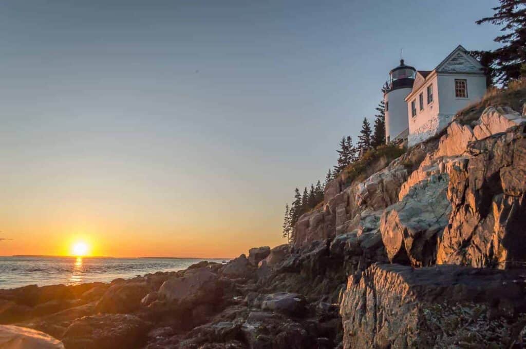 White lighthouse and building sit on a rocky cliff face against the water. The sun is setting in the distance at a popular tourist attraction Acadia National Park lighthouse.