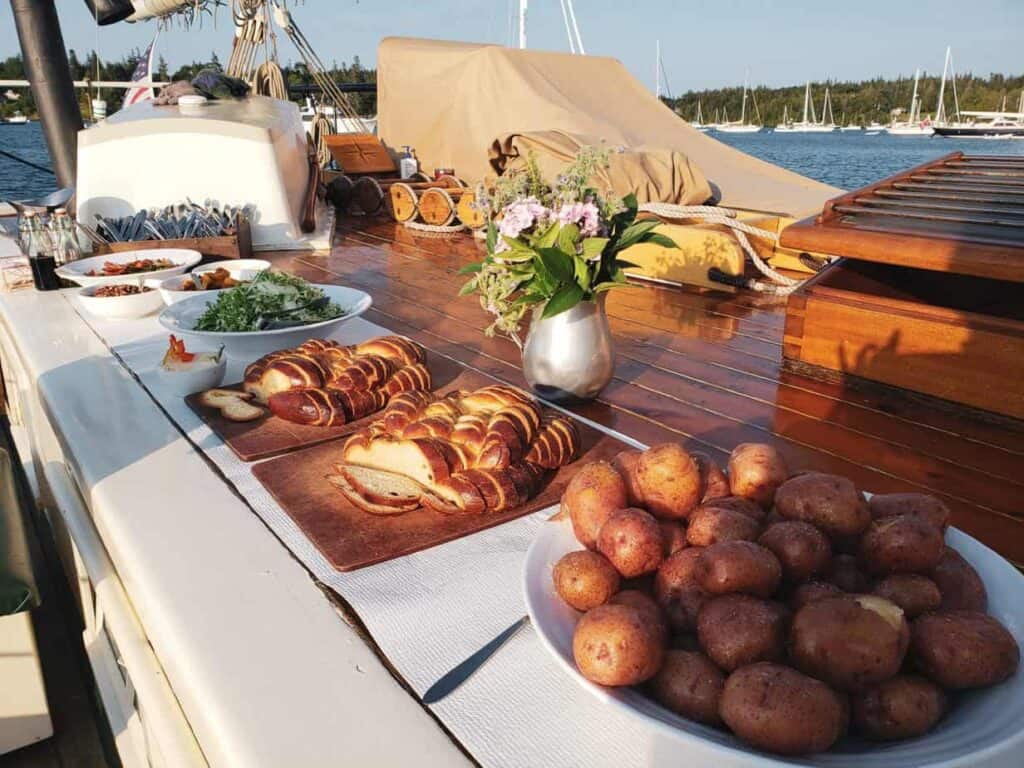 Foot sitting on a wooden table, featuring bread, potatoes, and salad. Boats floating on the water can be seen in the distance.