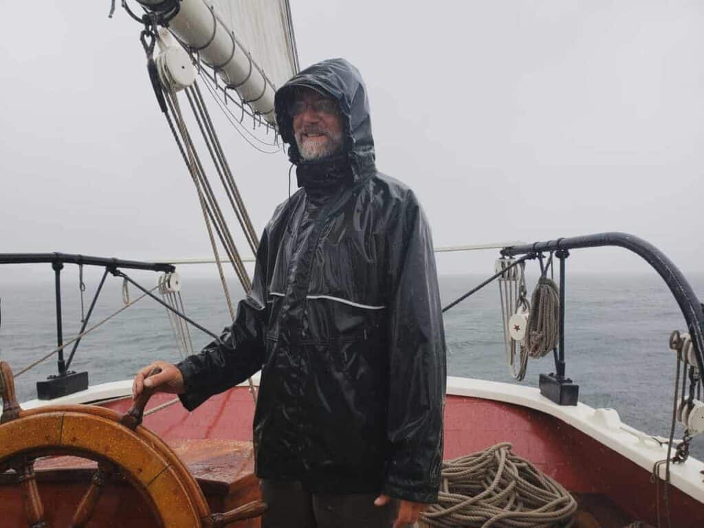 Man standing in a rain jacket on a boat surrounded by the water.