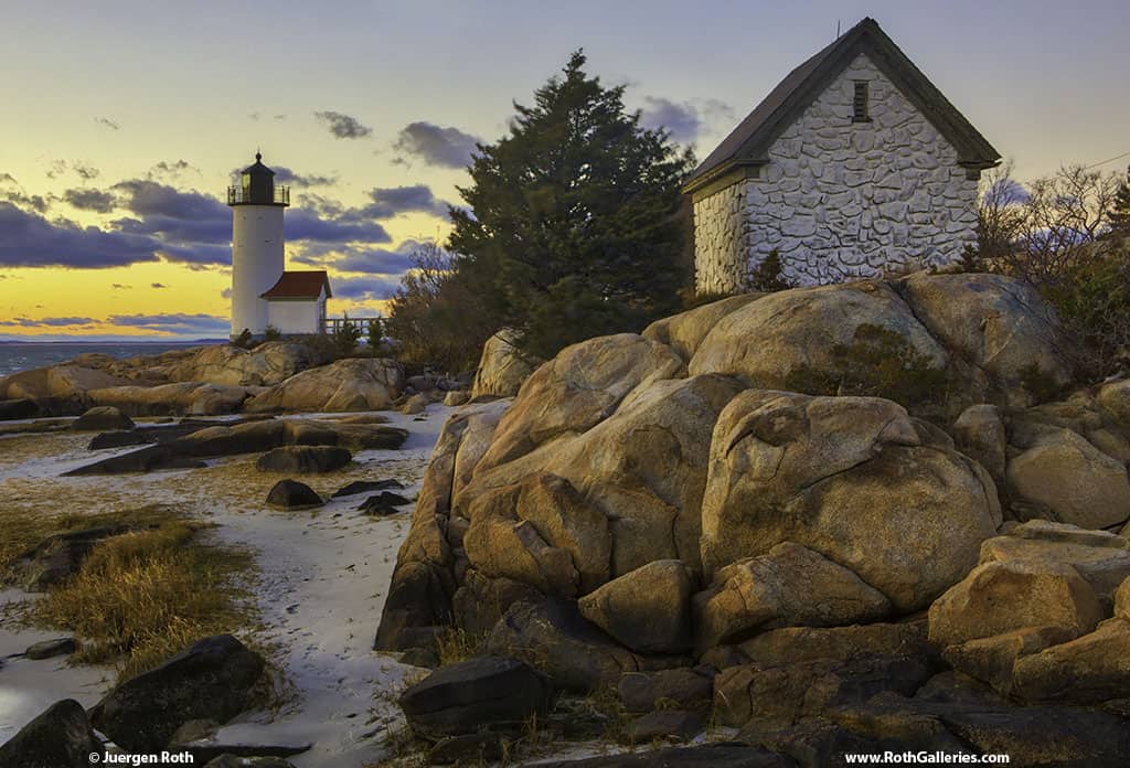 A rocky home is sitting on top of a boulder by the coast. In the distance, a white lighthouse is perched on the coast under a sunset sky.