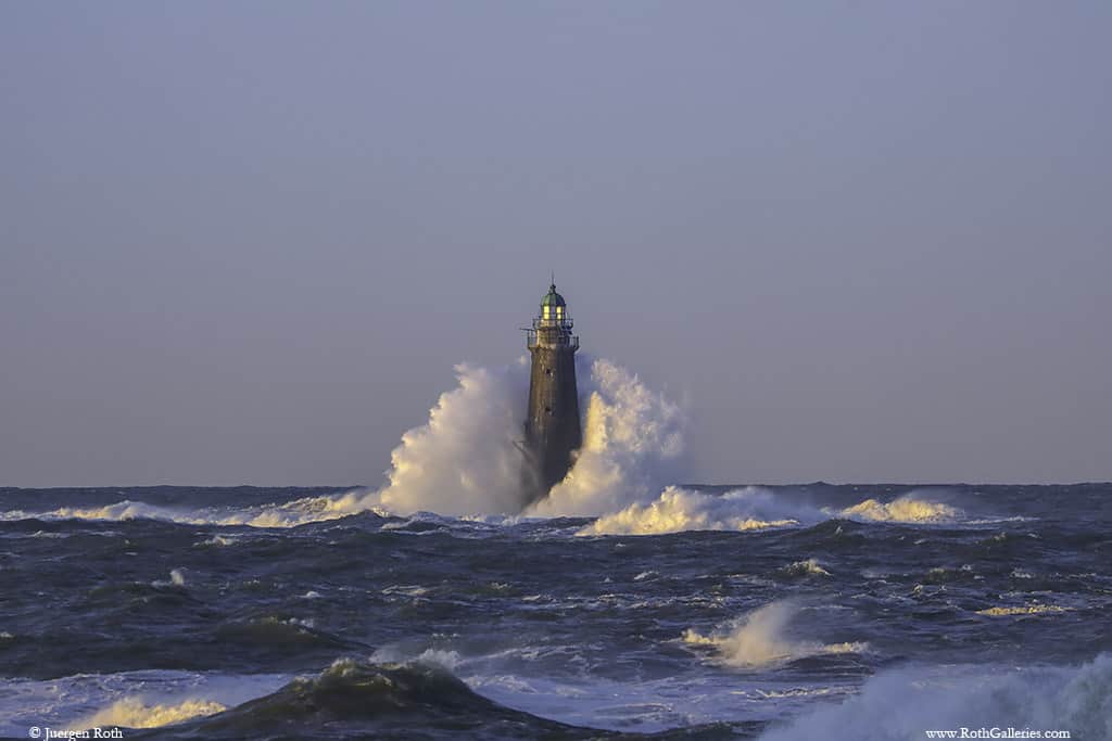 A lighthouse in the middle of the ocean has waves crashing around it.