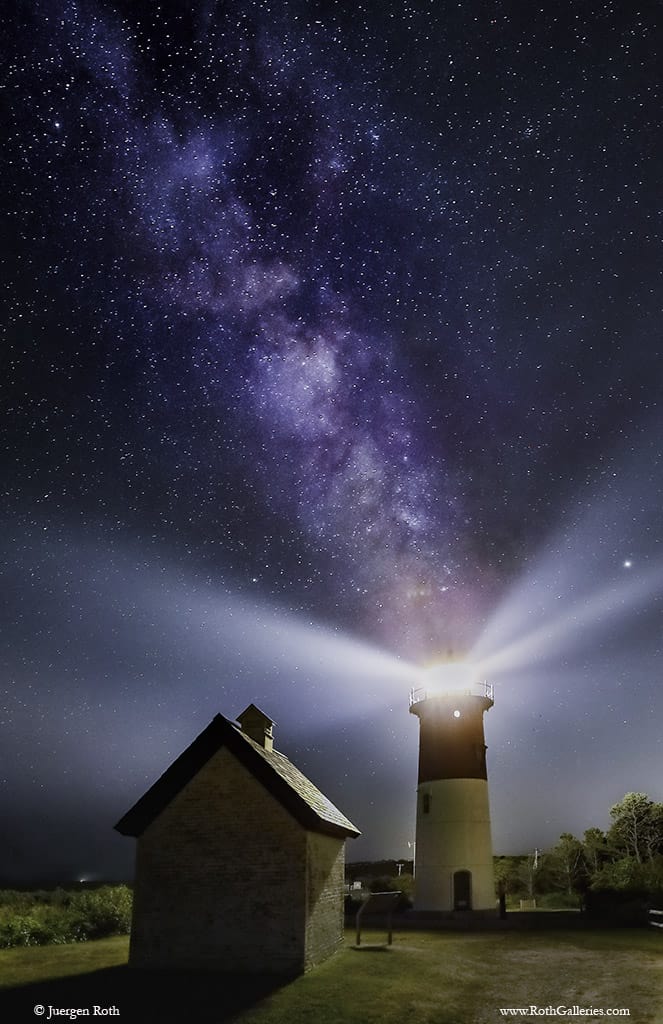 A lighthouse shines its lights under a starry sky next to a home at night.