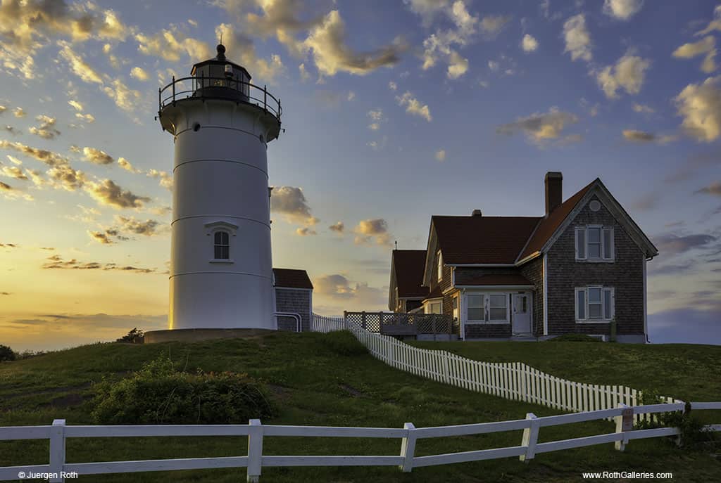 A white lighthouse is next to a historic red home surrounded by a white picket fence under a sunset.