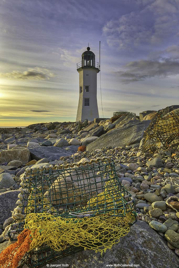 A white lighthouse is on a rocky coast under a sunrise.
