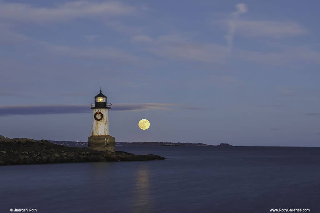An old black and white lighthouse with a holiday wreath sits on an island surrounded by water. The moon can be seen in the blue sky behind it.
