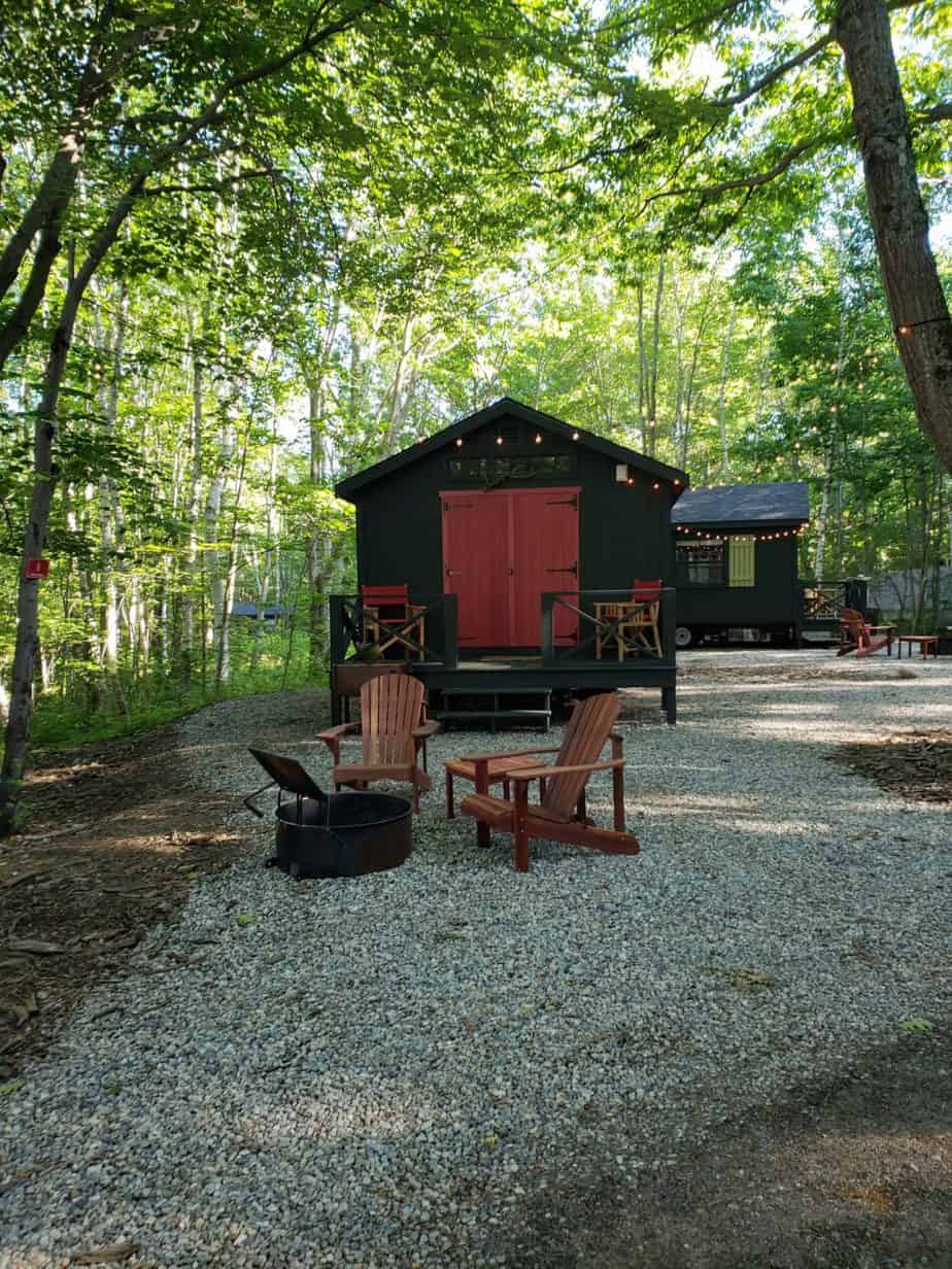 Small tiny brown cabin with a red door surrounded by forest. Fairy lights are on the front of the cabin. In the forefront, two outdoor chairs sit around a firepit.