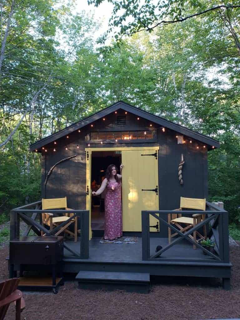A woman in a pink dress is opening the yellow doors of a dark brown cabin in the woods.