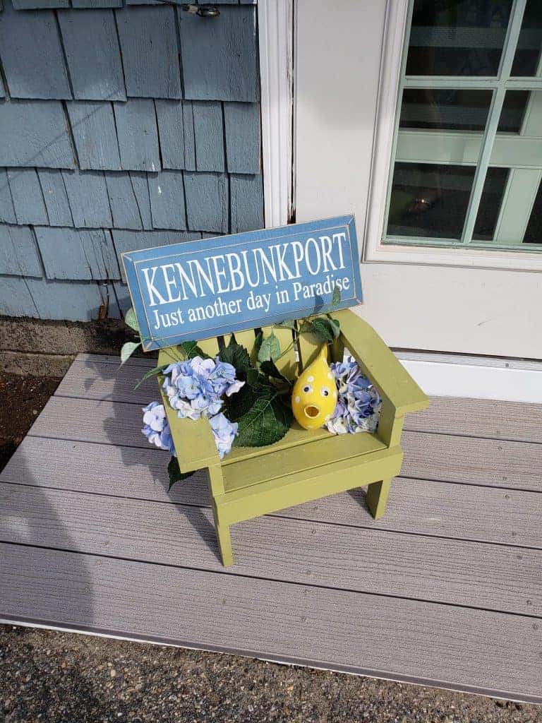 Blue sign with white writing that says \"Kennebunkport. Just another day in Paradise.\" on a yellow chair with flowers.