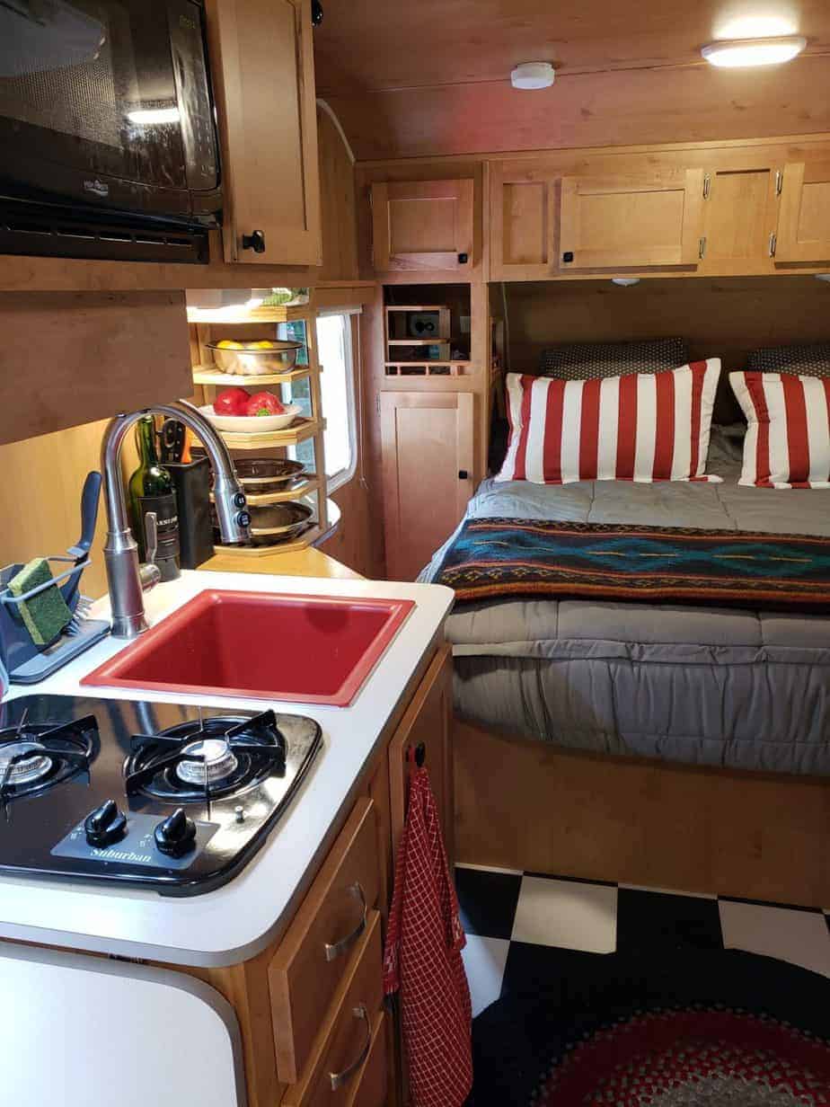 A stove top oven sitting inside of a kitchen with a red sink. A black and white checkerboard floor is seen. To the right, a bed is made with grey bedding and red and white striped pillows.