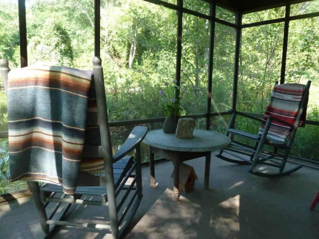 A screened porch with 2 rocking chairs and a table surrounded by green trees