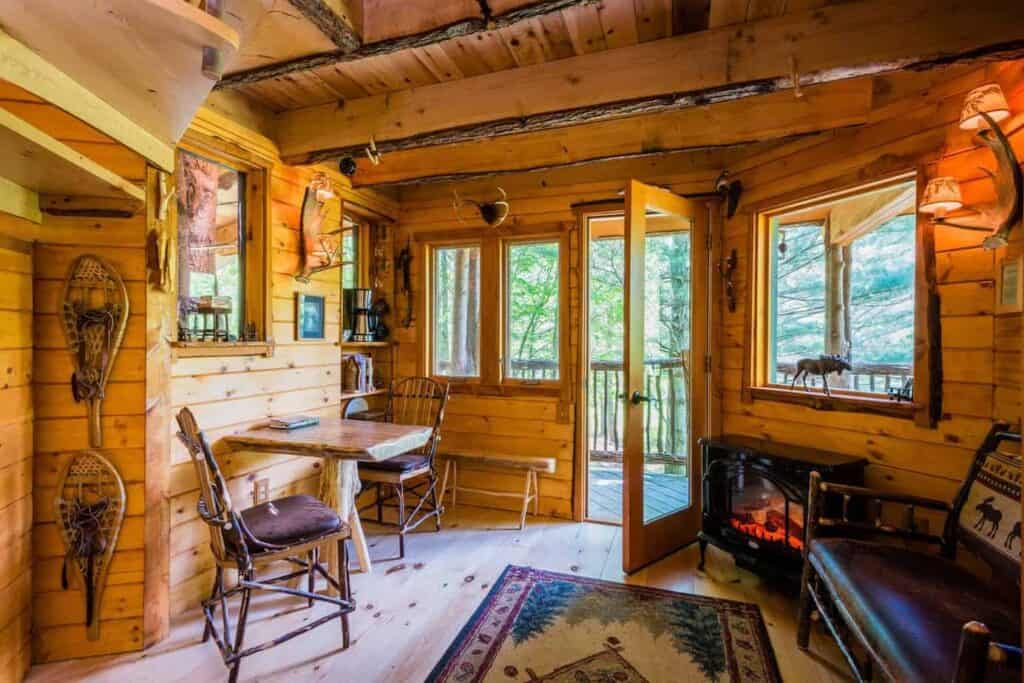 An open door in a wood paneled room with a table and chairs, rug and fireplace in a treehouse in Vermont