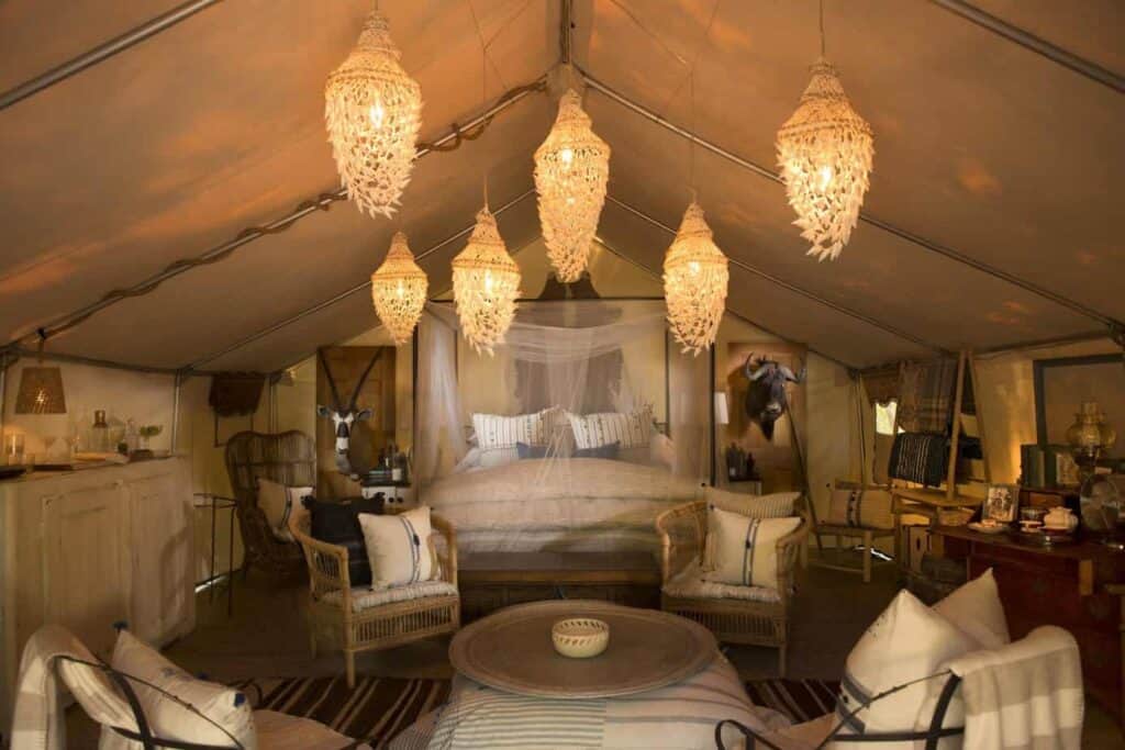 Interior of a glamping tent with cream tarp. A bed is made with a canopy shade around it to protect from bugs. Lights hang from the ceiling. Four wicker chairs sit around a coffee table in the forefront.