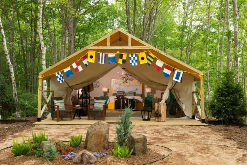 Exterior of a glamping tent with cream tarp surrounded by a green forest. Flags line the front of the tent.