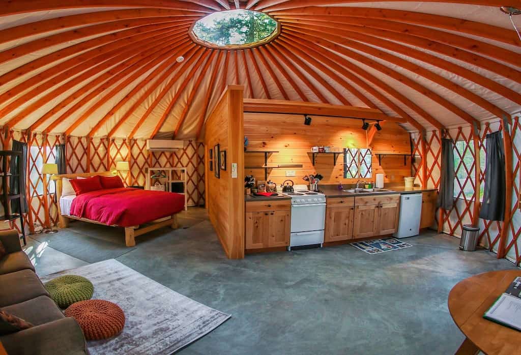 Interior of a yurt with a canopy ceiling. Inside, is a red bed, a sitting area, and a kitchenette.