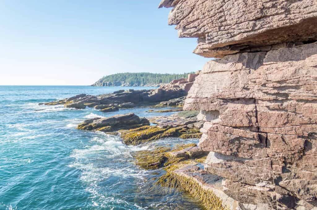 A rocky, dramatic, cliff with bright blue water under a blue sky is seen with a forest in the background.