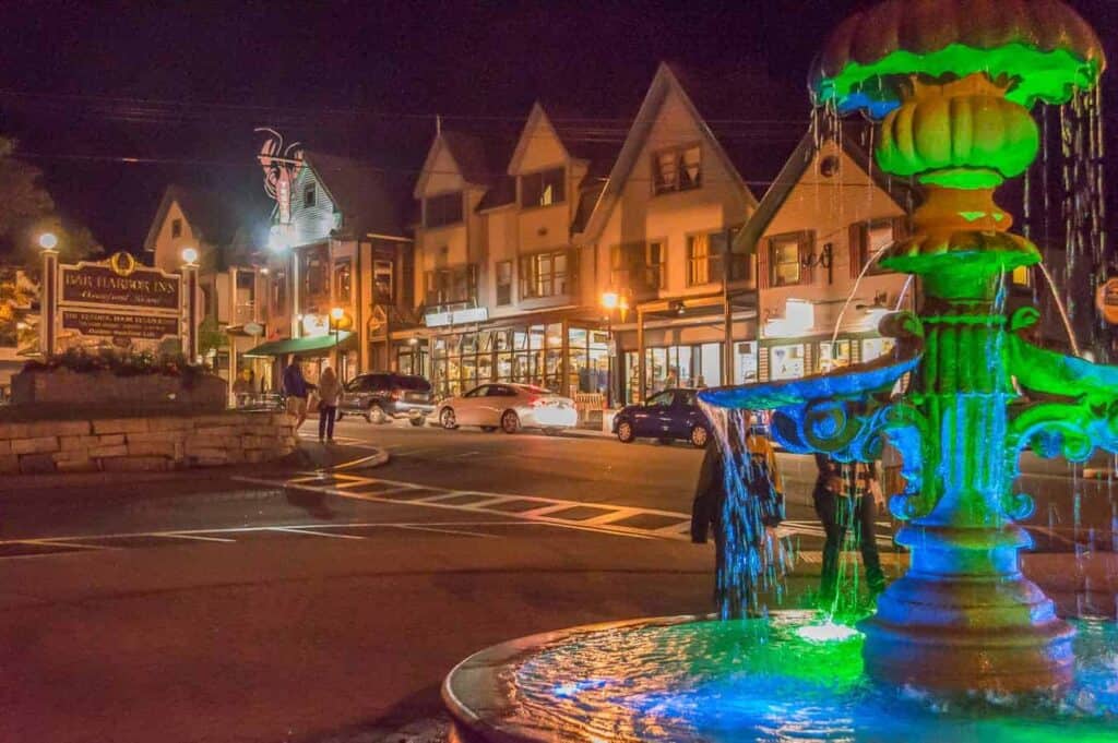 In the foreground, a fountain is lit up with green and blue lights. The background showcases shops lining a busy street with cars at night.