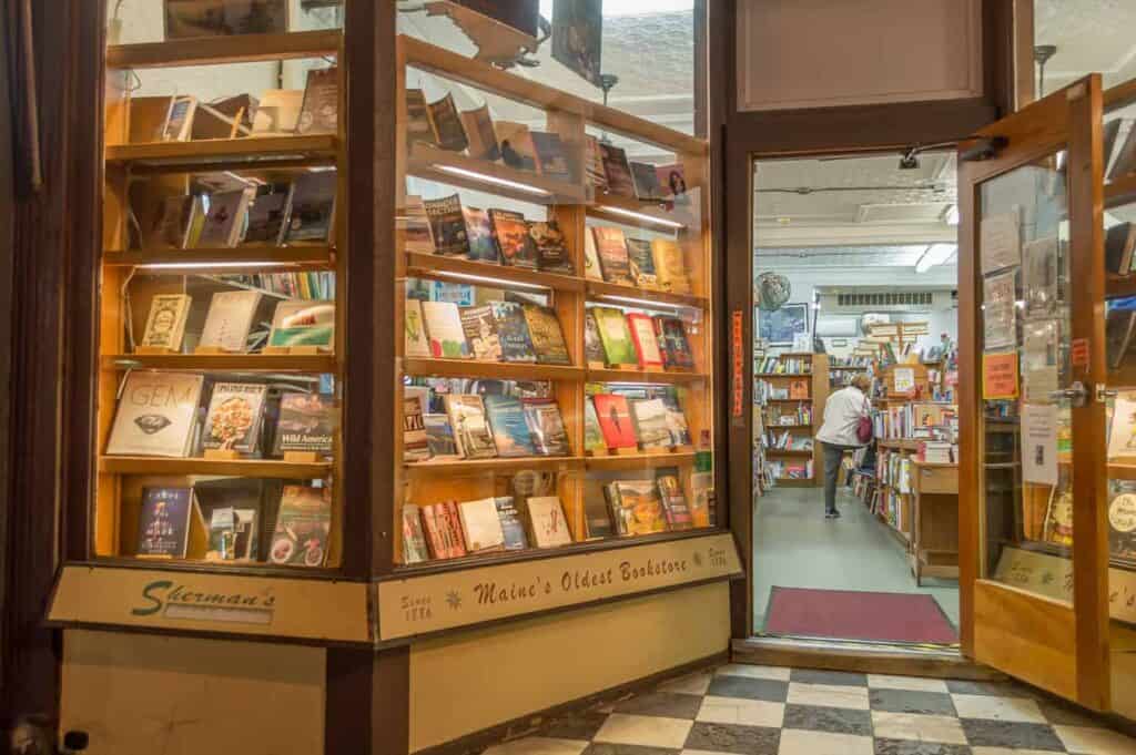 Entrance to a charming bookshop. The window showcases books on shelves, and a person can be seen through the window shopping.
