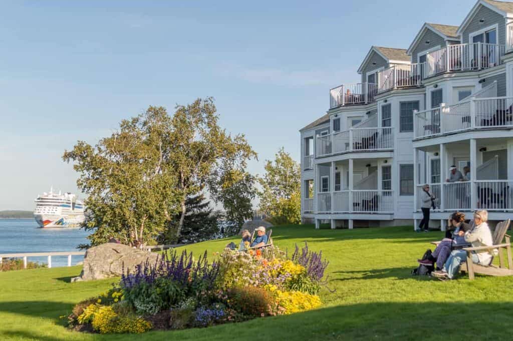 People sit on a lawn in comfortable chairs enjoying the weather. A white historic inn is seen in the distance under a blue sky.