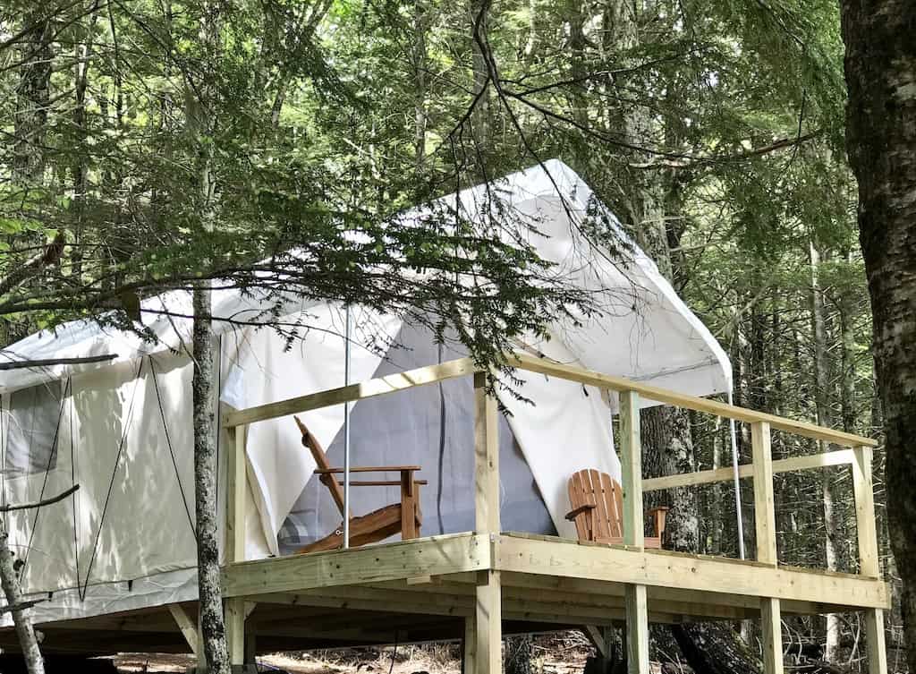 Exterior of a white tent with a deck, where two chairs are sitting, in the forest.