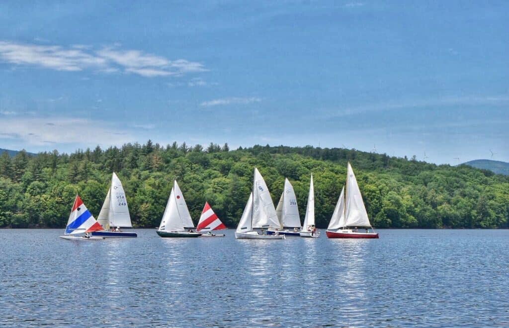 Several sailboats sail across a deep blue lake with green forest behind it and blue sky overhead