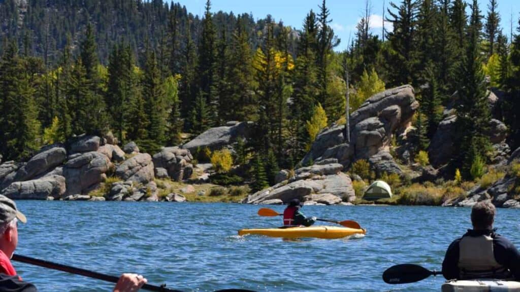 People are kayaking down a blue river with large boulders and evergreen forest on the bank