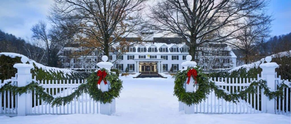 An opening in a fence leads to a white inn in the snow by trees. Two green wreaths hang off each side of the fence.