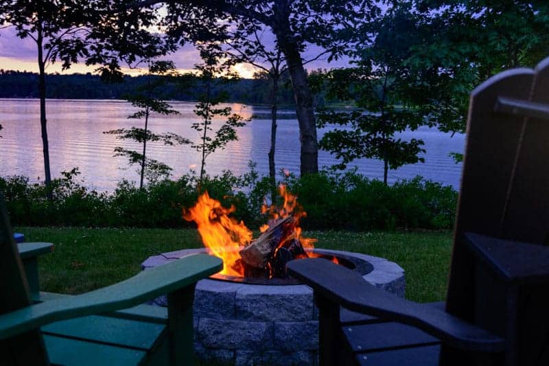 A fire burning in a fire pit in front of trees and a body of water at dusk