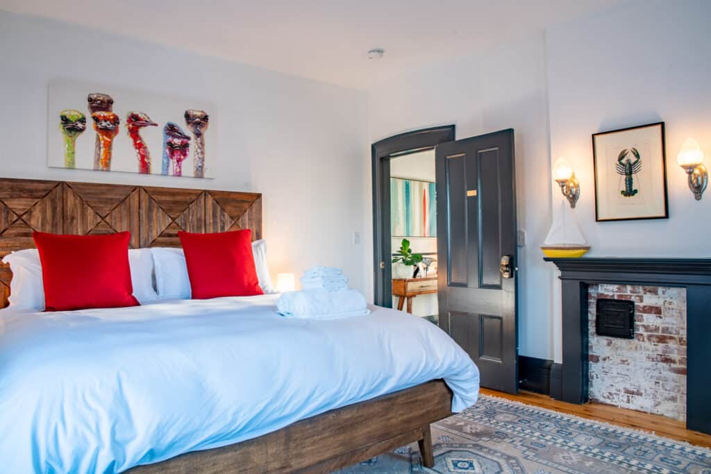 Wooden bed with a white soft comforter and red pillows. The slate grey door is open showing a table with plants in the next room during a Massachusetts couple trip