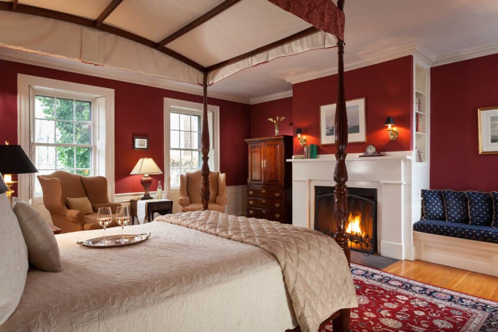 Interior of a romantic bedroom in a hotel with a fireplace lit. The walls are red, and there\'s a canopy bed as the centerpiece, with two glasses of champagne.